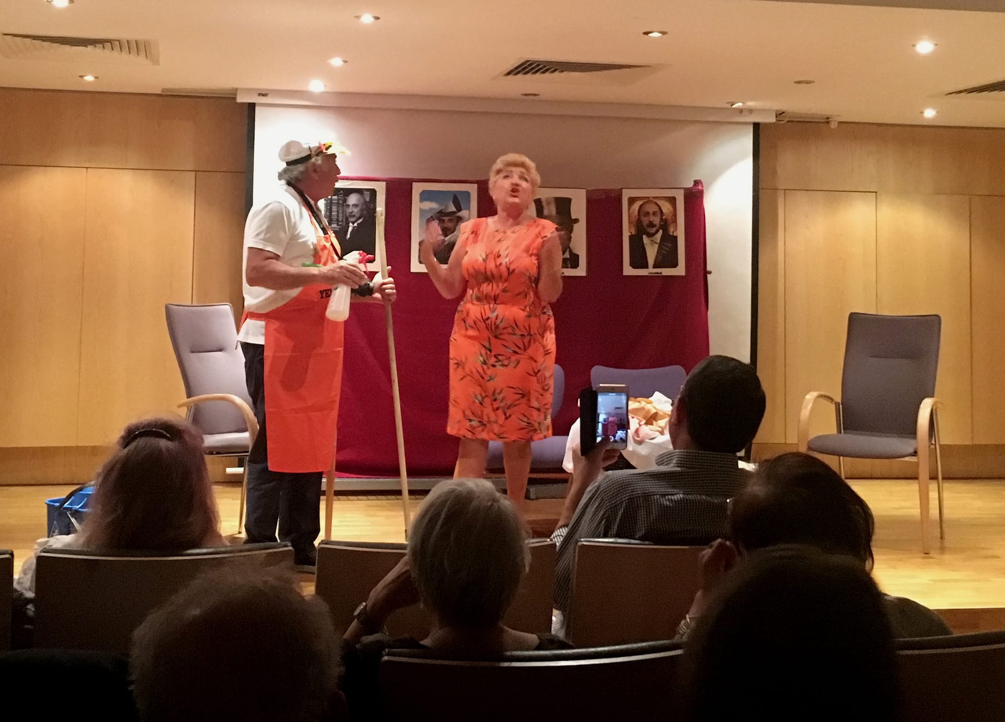 Onstage, a middle-aged woman in a pink floral dress looks upset and an older man in a cap and red apron looks on